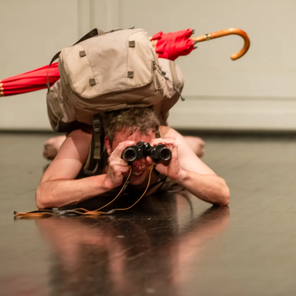 Performer Rok Kravanja na tleh z oprtanim nahrbtnikom, nekaj prav zadovoljno opazuje skozi daljnogled.