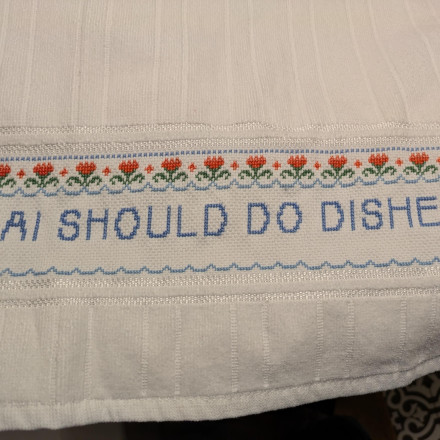Bela kuhinjska krpa z vezenino - vrstico rožic z rdečimi cvetovi in modrim napisom "AI SHOULD DO DISHES"